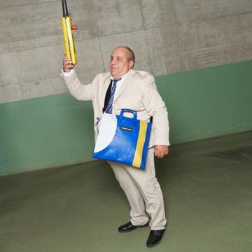 A man wearing a cream suit, white shirt and blue striped tie, with a blue, white and yellow truck-tarp FREITAG bag worn cross-body, reaches up to a yellow crane controller in a concrete-and-green factory hall.