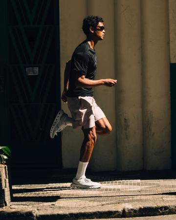 A man wearing sunglasses, a black short-sleeve performance tee, mauve running shorts, white crew socks and white On running shoes, caught mid-stride on a sunlit mosaic pavement beside a pale fluted wall.