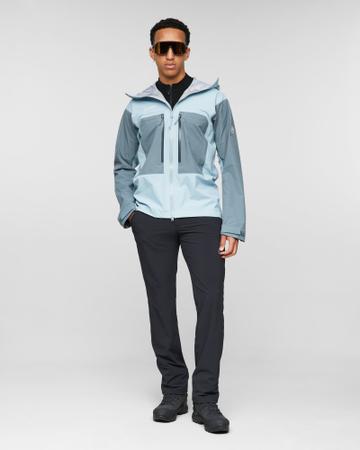 A man wearing a zipped light-blue and slate-grey Mammut Taiss HS hooded hardshell jacket over a black half-zip top, with black trekking trousers, wraparound sunglasses and dark hiking boots, standing full length against a grey studio background.