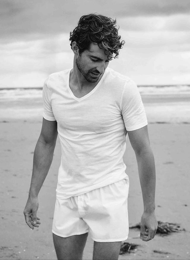 A man walking on a beach in a white Hanro cotton V-neck T-shirt and white boxer shorts, in black and white.