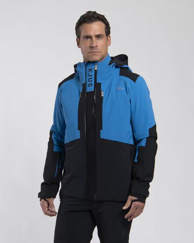 A man wearing a cobalt-blue and black Kjus hooded ski jacket with KJUS printed down the collar, zipped to the chin, and black ski trousers, on a pale grey studio background.
