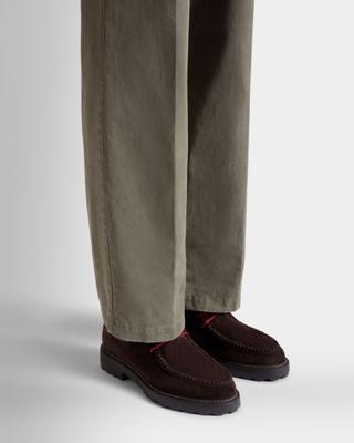 A man wearing wide olive cotton trousers over dark brown suede moc-toe derby shoes with red laces and chunky lug soles, shown from the knee down on a grey studio floor.