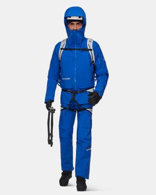 A man wearing a cobalt-blue Mammut hooded hardshell jacket and matching shell pants, with the hood up over a white helmet, a climbing harness, a light grey backpack, black gloves and black boots, holding an ice axe in his right hand against a light grey studio background.
