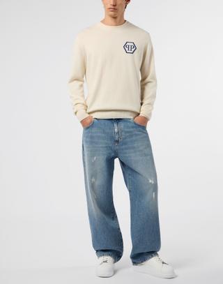 A man wearing a cream knit crewneck sweater with a navy hexagon PP logo on the chest, tucked slightly at the front of baggy light-wash wide-leg jeans with distressed details, and white sneakers, standing with his hands in his pockets against a pale grey studio background.
