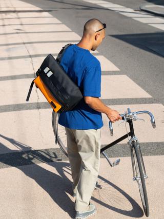 A man wearing a cobalt-blue tee, stone chinos and a black-and-orange messenger bag slung cross-body on his back, holding a black road bike with chrome handlebars and fork on a sunlit zebra crossing, seen from behind at a high angle.