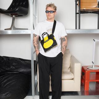A man wearing blue-lens sunglasses, a white graphic tee with a daisy print, and wide black pleated trousers, carrying a small black-and-yellow FREITAG truck-tarp crossbody bag across his chest, standing hands in pockets in front of metal shelving holding vintage chairs.