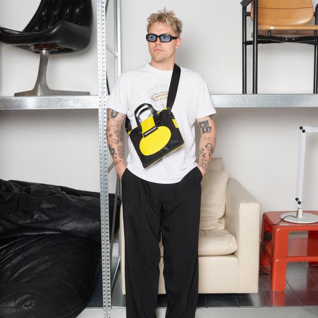 A man wearing blue-lens sunglasses, a white graphic tee with a daisy print, and wide black pleated trousers, carrying a small black-and-yellow FREITAG truck-tarp crossbody bag across his chest, standing hands in pockets in front of metal shelving holding vintage chairs.