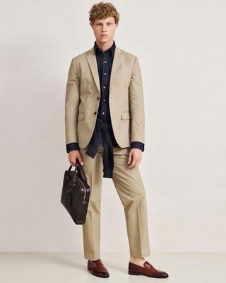 A man wearing a light-beige cotton two-button suit over a dark denim shirt, with a navy sweater knotted at the waist, burgundy leather buckle loafers, and a dark brown leather briefcase in his right hand, standing full length against a pale studio background.