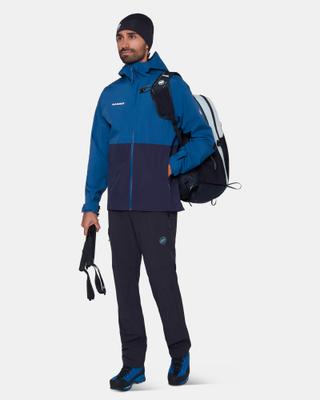 A bearded man wearing a two-tone blue and navy Mammut hooded hardshell jacket, black Mammut trekking trousers, a black beanie and blue mountaineering boots, with a backpack slung over one shoulder and folded trekking poles in his hand, standing full length on a pale studio background.