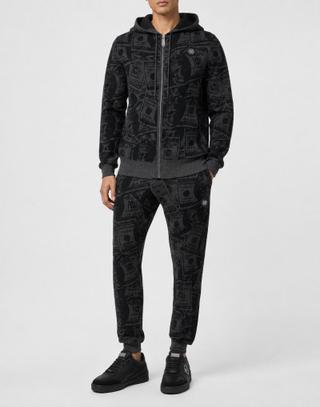 A man wearing a charcoal-and-black all-over dollar-bill jacquard knit zip hoodie with ribbed hem and cuffs, matching cuffed joggers with a logo patch, and black leather logo sneakers, standing against a pale grey studio background.