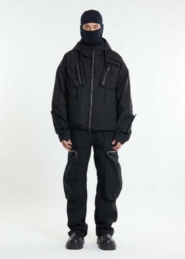 A man wearing a black hooded padded jacket zipped to the chin with zipped chest pockets and dangling toggle cords, a navy balaclava, black cargo trousers with curved zip thigh pockets, and chunky black leather boots, standing in a white studio.