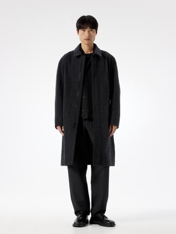 A man wearing a knee-length charcoal wool coat open over a black shirt, straight black trousers, and black leather derby shoes, standing in a white studio.