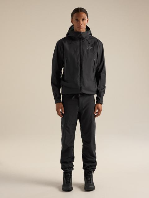 A man wearing a black Arc'teryx hooded shell jacket zipped to the chin, slim black technical trousers gathered at the ankle, and black hiking boots, standing on a beige studio background.