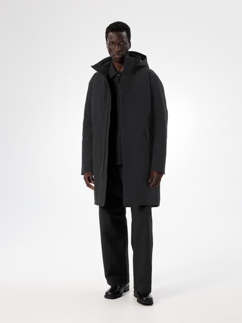 A man wearing a black hooded knee-length down coat with a smooth shell face, worn open over a black zip-neck top, with straight black trousers and black leather shoes, standing head to toe in a white studio.