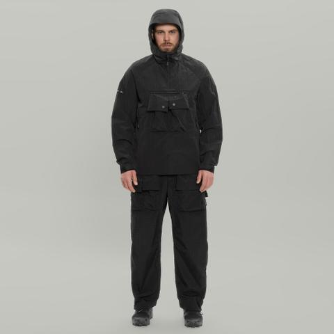 A man wearing a black crinkle-nylon anorak with the hood up and a large kangaroo chest pocket, black ripstop cargo trousers with flap pockets, and black trail sneakers, standing against a plain light-grey background.
