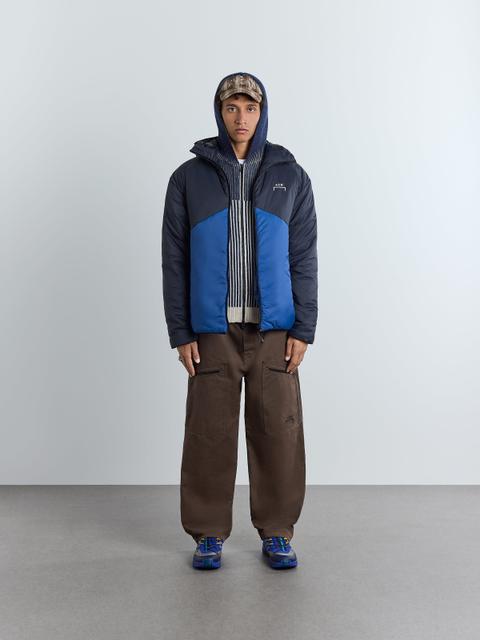 A man wearing a navy and cobalt-blue hooded insulated A-COLD-WALL jacket open over a striped hooded zip knit, wide brown trousers with zipped thigh pockets, blue technical trail sneakers and a check cap, standing in a studio.