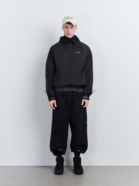 A man wearing a black hooded A-COLD-WALL nylon jacket with toggle drawcords and a chest logo, zipped over a black roll neck, with black cuffed sweatpants, black hiking boots and a cream logo cap.