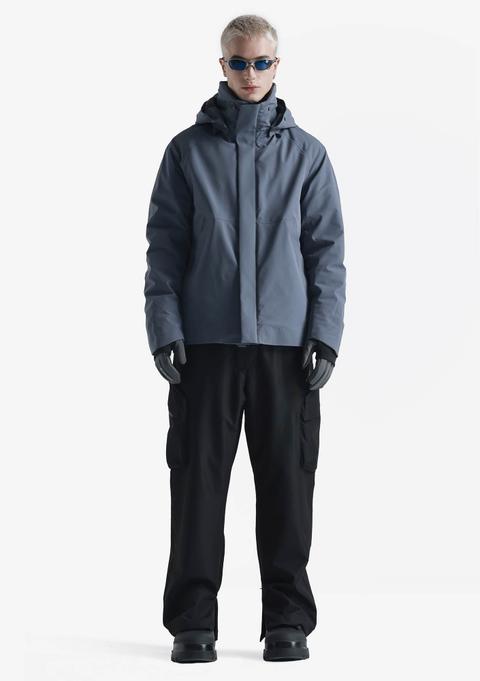 A man wearing a slate-blue hooded insulated technical jacket zipped up to a high storm collar, grey gloves, wraparound blue sunglasses, wide black cargo trousers and chunky black boots, standing against a white studio background.