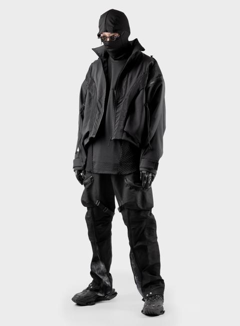 A man wearing a cropped black technical jacket open over a layered black mesh-panel top, with a black balaclava, round goggles, black gloves, black articulated cargo trousers with buckled thigh straps and zipped pocket bags, and chunky black sneakers, standing in a white studio.