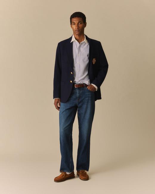 A man wearing a navy two-button blazer with gold buttons, patch pockets and a crest, worn open over a blue-and-white striped oxford shirt with a white collar, a brown leather belt, mid-wash straight jeans, white socks and tan suede boat shoes, standing on a plain beige studio background.
