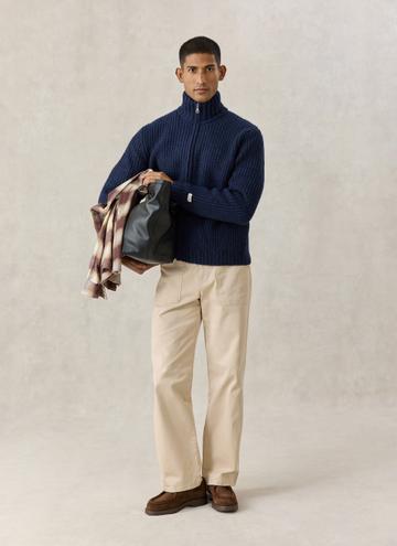 A man wearing a navy ribbed zip-neck fisherman cardigan, stone patch-pocket chinos and brown suede moc-toe shoes, carrying a black leather tote and a brown check overshirt over his arm, standing on a plain stone studio ground.