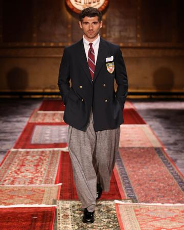 A man wearing a navy double-breasted blazer with gold buttons, a crest patch and a white pocket square over a white shirt and burgundy rep-stripe tie, with wide pleated grey tweed trousers and black loafers, walking a runway lined with Persian rugs.