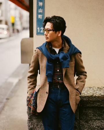 A man wearing tortoiseshell glasses, a brown herringbone tweed sport coat over a red-and-navy tartan button-down and white tee, a navy sweatshirt knotted over his shoulders, a dark belt and dark indigo jeans, standing on a Tokyo street beside a Kamiyamacho sign and holding a paisley scarf.