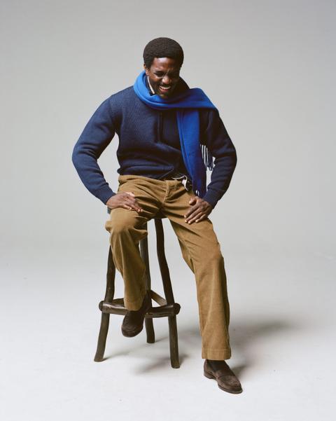 A man wearing a navy knit sweater over a collared shirt, a bright blue wool scarf, tan corduroy trousers, dark socks and brown suede penny loafers, seated on a wooden stool against a plain grey studio backdrop.