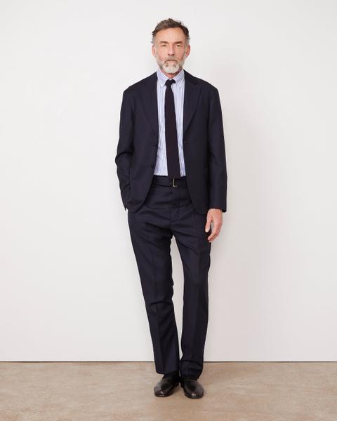 A man wearing a navy wool two-button suit with the jacket open, a blue-and-white striped button-down collar shirt, a navy knit tie, a dark belt, navy socks and black leather slip-on loafers, standing against a plain white wall.