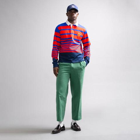 A man wearing a red-and-blue striped long-sleeve rugby shirt with a white collar and navy hem, pleated green cropped chinos, a navy NY cap, white socks and dark burgundy penny loafers, standing on a plain grey studio background.
