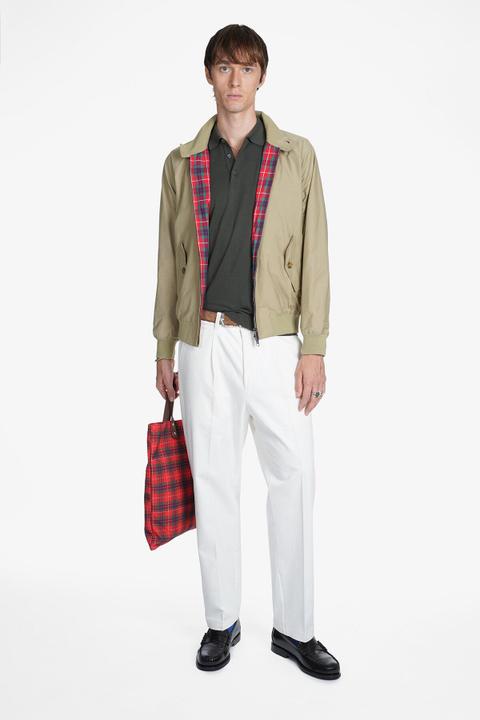 A man wearing a stone Harrington jacket open to show its red tartan lining over a dark olive knit polo, pleated off-white trousers, blue socks and black penny loafers, holding a red tartan tote bag against a plain white background.