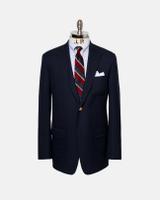 A navy single-breasted blazer displayed on a J. Press dress form, front view, with a gold button fastened at the waist, flapped patch hip pockets, and a patch chest pocket with a white pocket square, worn over a blue-and-white striped button-down shirt and a navy, red, and gold striped rep tie.