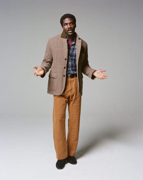 A man wearing a brown herringbone tweed games blazer with a cord collar over a blue plaid flannel shirt and burgundy tee, tan double-knee canvas trousers, a black belt and black suede loafers, standing on a plain grey studio ground.
