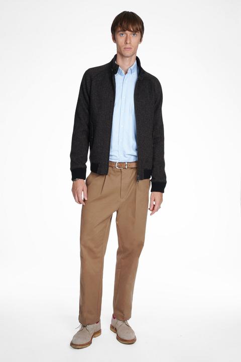 A man wearing a dark brown herringbone wool Harrington jacket open over a light blue oxford button-down shirt, pleated tan chinos with a tan suede belt, and sand suede desert boots, standing on a plain white background.