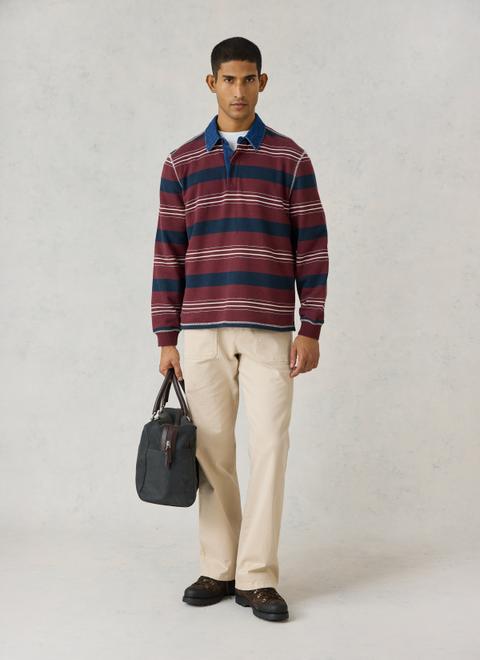 A man wearing a burgundy-and-navy striped rugby shirt with a denim collar over a white tee, stone patch-pocket chinos and brown lace-up hiking boots, carrying a charcoal holdall.