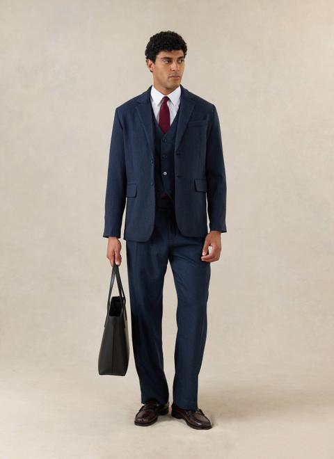 A man wearing a navy herringbone two-button jacket with matching waistcoat and trousers, a white point-collar shirt, a burgundy knit tie and burgundy penny loafers, holding a black leather tote, standing on a plain stone studio ground.