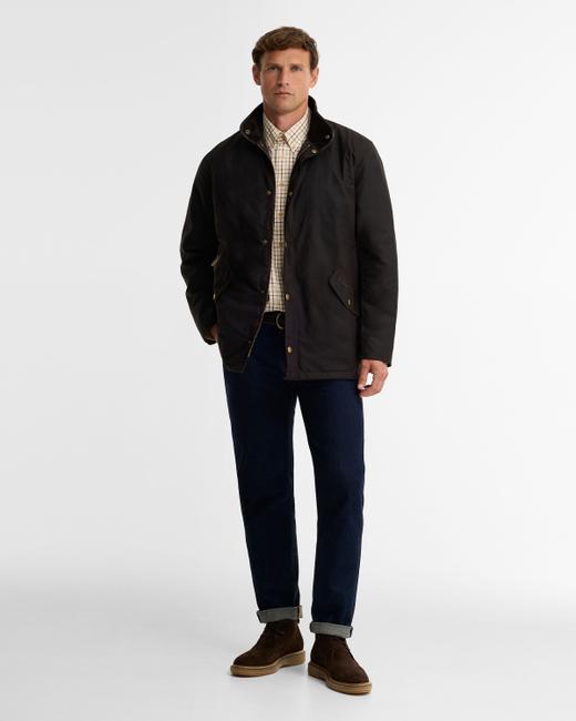 A man wearing a dark brown waxed cotton jacket worn open with a dark cord collar, over a cream tattersall-check shirt and a dark belt, with dark rinse straight jeans finished with a single turn-up cuff and brown suede chukka boots with crepe soles, standing on a plain white studio background.