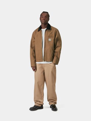 A man wearing a Hamilton-brown duck canvas Carhartt WIP Detroit Jacket with a black corduroy collar, worn open over a plain white tee, with relaxed khaki twill work pants and black leather lace-up derby shoes, standing on a plain pale studio background.