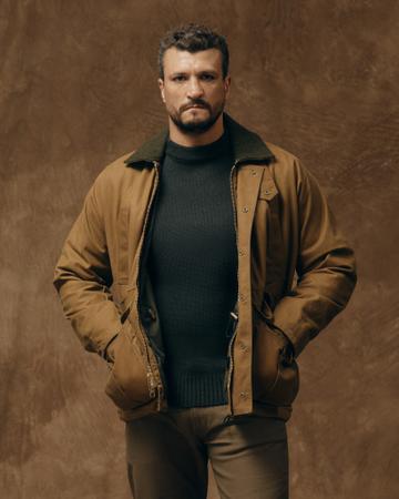A man wearing a dark-tan Filson Lined Tin Cloth Field Jacket with an olive wool-lined collar, worn open over a dark green crew-neck wool sweater with brown trousers, hands in the jacket pockets, against a mottled brown backdrop.