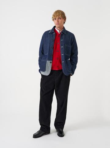 A man wearing an open indigo denim chore jacket with a contrast chambray lower patch pocket over a red wool cardigan and white collared shirt, with relaxed black corduroy pleated trousers and black leather penny loafers, standing on a plain white studio background.