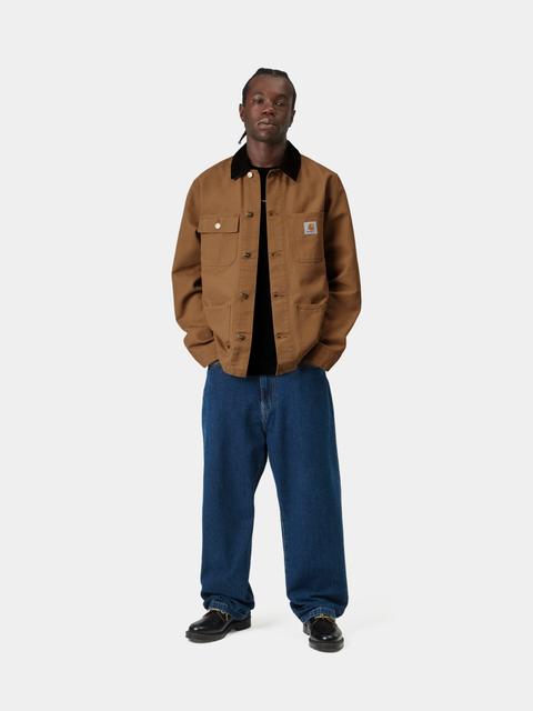 A man wearing a brown duck canvas chore coat with a black corduroy collar, open over a black tee, with wide dark-blue jeans and black leather lace-up shoes, standing on a pale grey studio background.