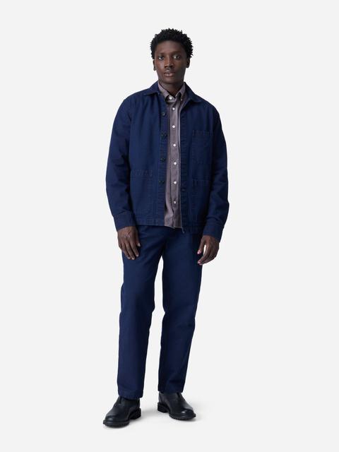 A man wearing an open indigo cotton chore jacket with three patch pockets over a mauve-grey button-down shirt, matching indigo straight work trousers and black leather lug-sole boots, standing on a plain light-grey studio background.