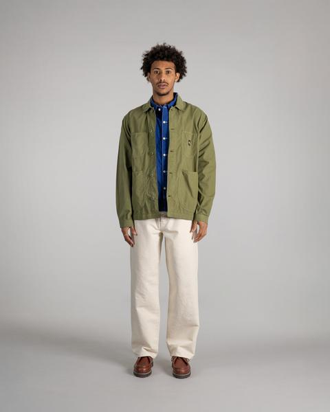 A man wearing an unbuttoned olive cotton coverall jacket with patch pockets over a cobalt-blue work shirt, wide ecru trousers and brown leather moc-toe shoes, standing on a plain grey studio background.