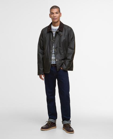 A man wearing a dark olive waxed jacket with a brown corduroy collar, worn open over a navy and cream patterned crew-neck jumper, with a dark leather belt, dark rinse straight jeans with turned-up cuffs and dark brown leather lace-up moc-toe boots, standing on a plain white background.