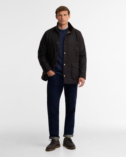 A man wearing a dark rustic-brown waxed cotton jacket with a corduroy collar, worn open over a navy marled crew-neck wool sweater with a check shirt collar showing, dark rinse straight jeans with a turn-up cuff and grey suede lace-up derby shoes, standing on a plain white studio background.