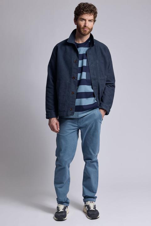 A man wearing a navy cotton fisherman work jacket with patch pockets, worn open over a navy and sky-blue wide-striped top, with straight washed-blue cotton trousers and navy-and-cream suede running sneakers, standing on a plain pale-grey studio background.