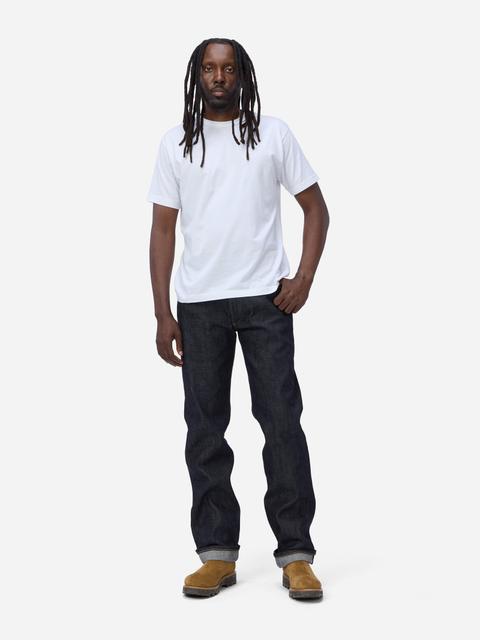 A man wearing a plain white crew-neck tee, dark raw indigo straight-leg selvedge jeans with a single cuff, and tan suede lug-sole boots, standing on a plain light-grey studio background.