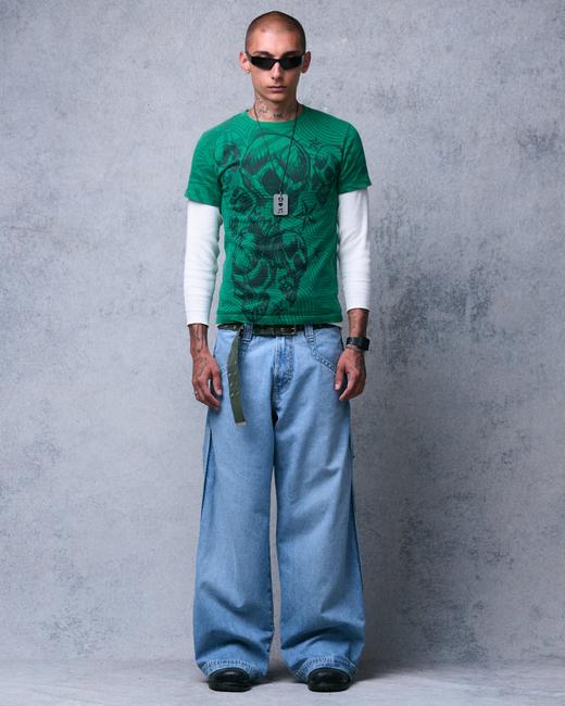 A man wearing wraparound black sunglasses, a dog-tag necklace, a green skull-print graphic tee layered over a white waffle long-sleeve, an olive grommet web belt with a hanging tail, and very wide-leg light-wash jeans pooling over black shoes, standing against a plain grey backdrop.