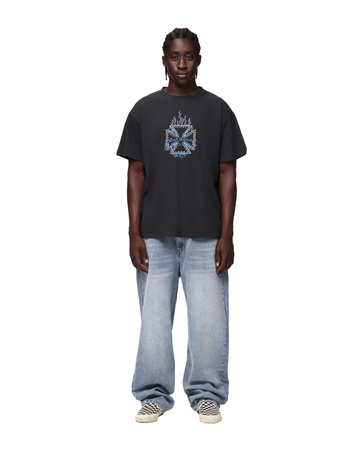 A man wearing a washed-black Von Dutch t-shirt with a studded iron-cross graphic and blue flames, loose light-wash straight-leg jeans and black-and-white checkerboard slip-on sneakers, standing on a white background.