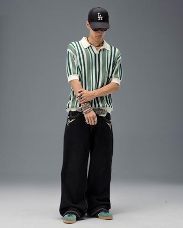 A man wearing a black LA cap, wraparound sunglasses, a green-and-cream vertically striped knit polo, stacked silver chain bracelets and rings, a studded western belt, very wide black JNCO jeans with cream tribal embroidery on the front pockets, and teal suede sneakers, standing on a grey studio backdrop.